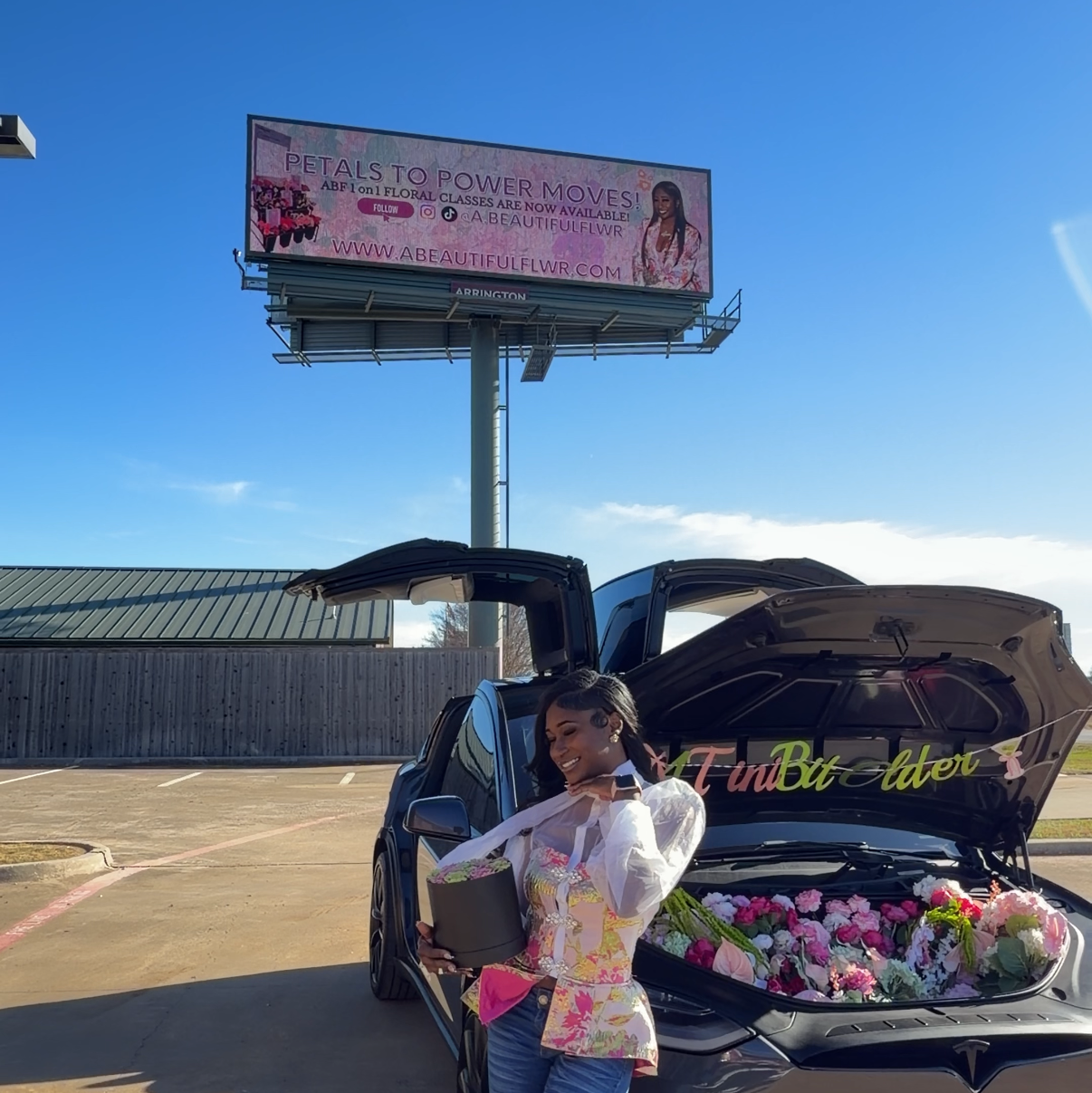 Woman standing next to a car with an open trunk full of flowers, in front of a billboard.